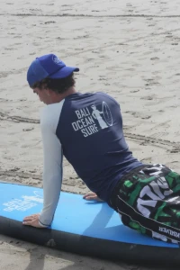 Surfing instructor wearing a navy blue Bali Ocean Surf long-sleeved rash guard and blue cap, lying on a surfboard in Bali