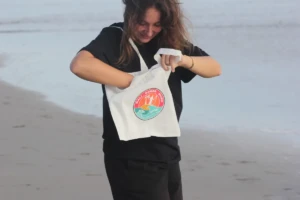 Young woman holding a white Bali Ocean Surf tote bag with a colorful sunset logo on Legian Beach, Bali.