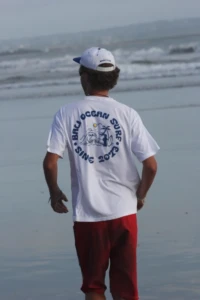 Man from behind wearing a white Bali Ocean Surf t-shirt with a blue "Since 2013" logo on the beach in Bali