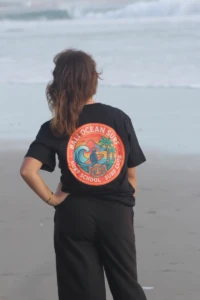 Woman from behind wearing a black Bali Ocean Surf t-shirt with a large, colorful logo Surf School Surf Club by the sea in Bali